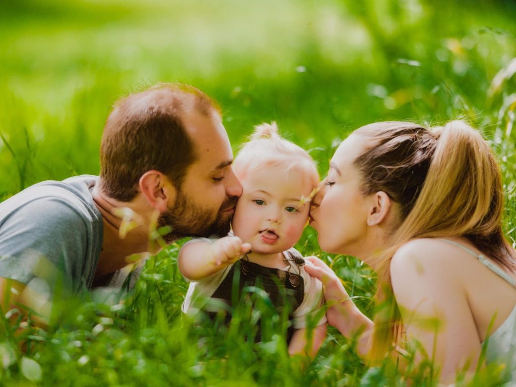 Photo of a small boy with his Dad kissing his left cheek and his mum kissing his right cheek,