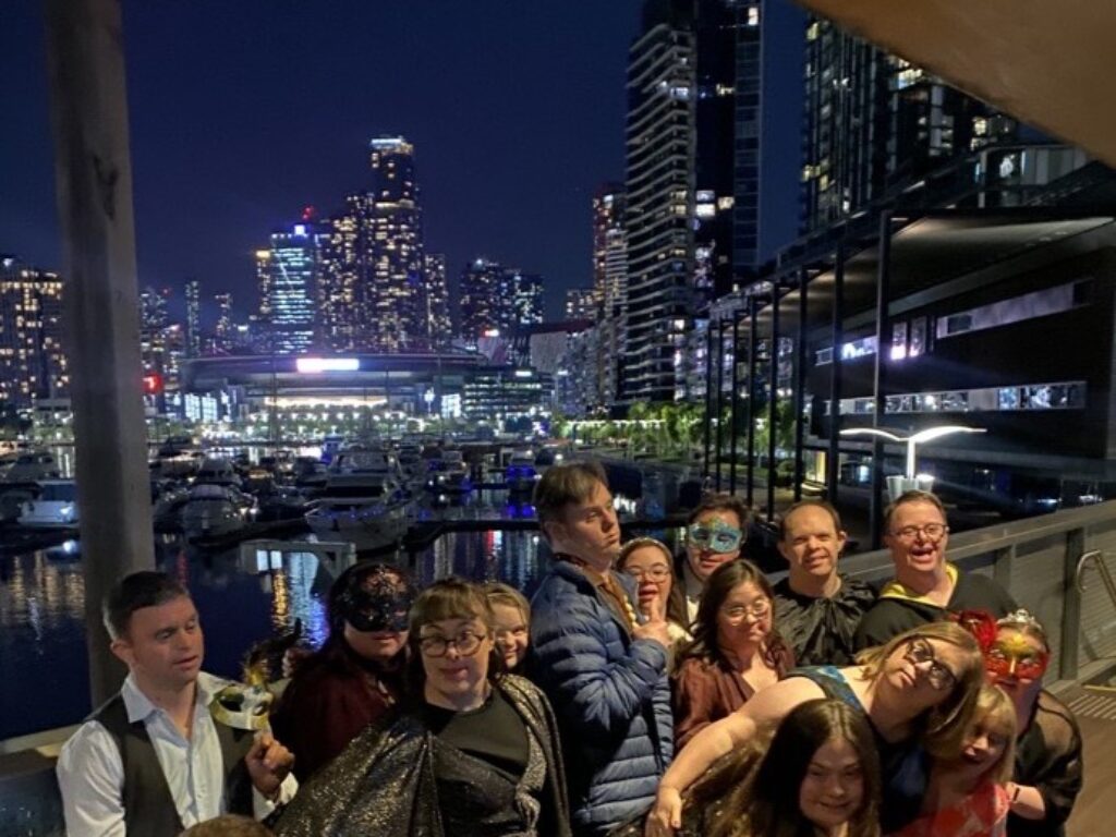 A group of adults with Down syndrome at the Club21 Masquerade winter ball. They are standing together on a balcony at nighttime. They are at Docklands with water and city buildings in the background.
