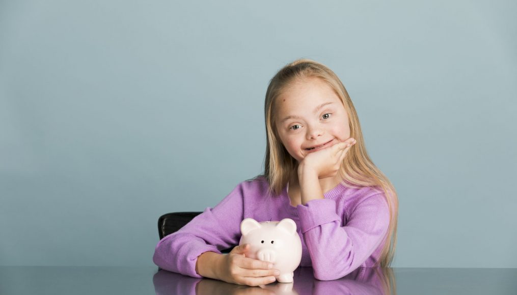 A young blonde girl with Down syndrome is seated at a table smiling at the camera and holding a piggy bank.