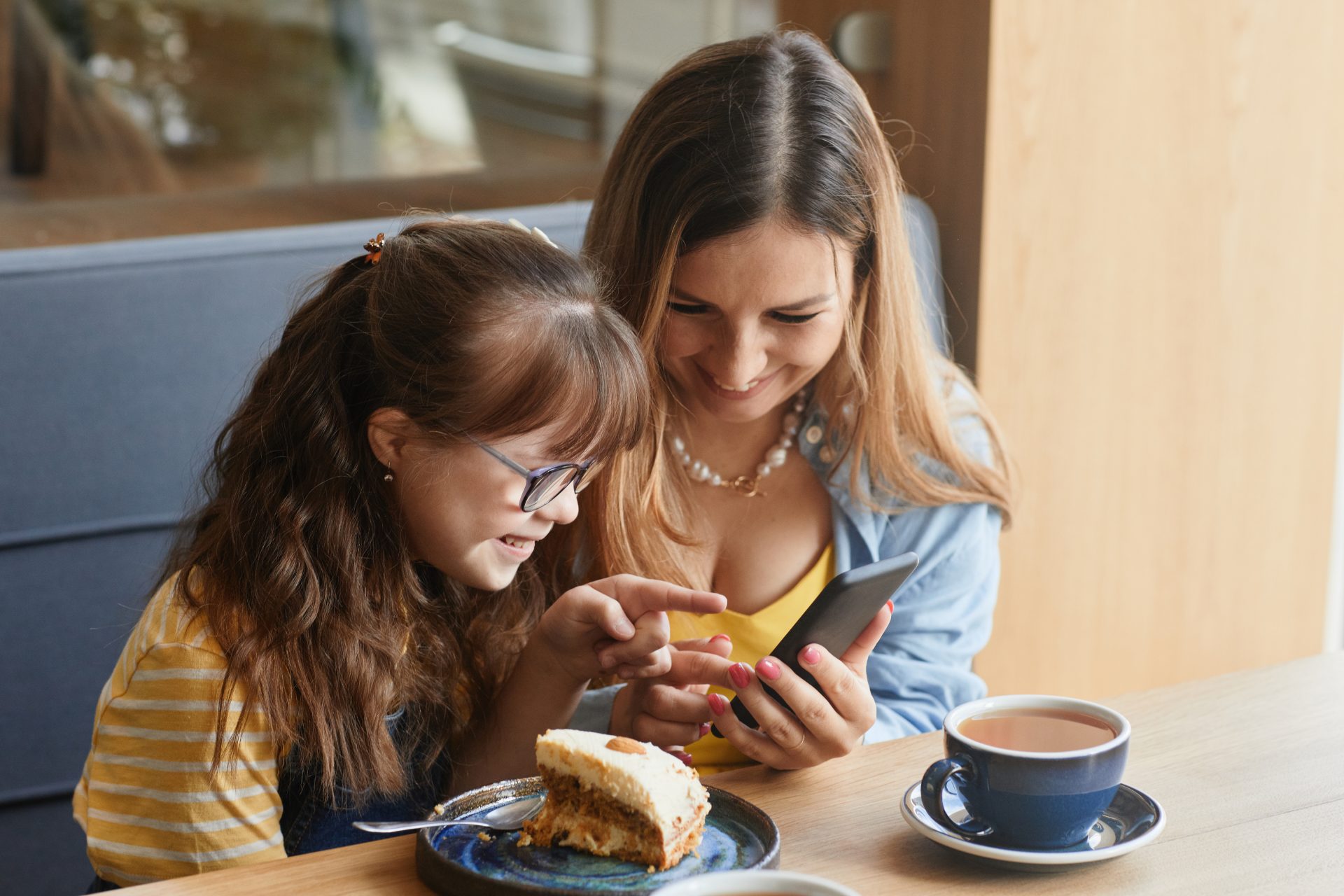 A woman and a young girl with Down syndrome are sitting in a cafe looking at a mobile phone screen together and smiling.