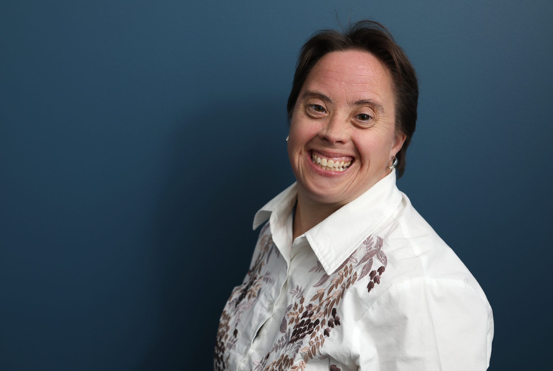 A woman with Down syndrome in a white collared shirt is smiling at the camera.