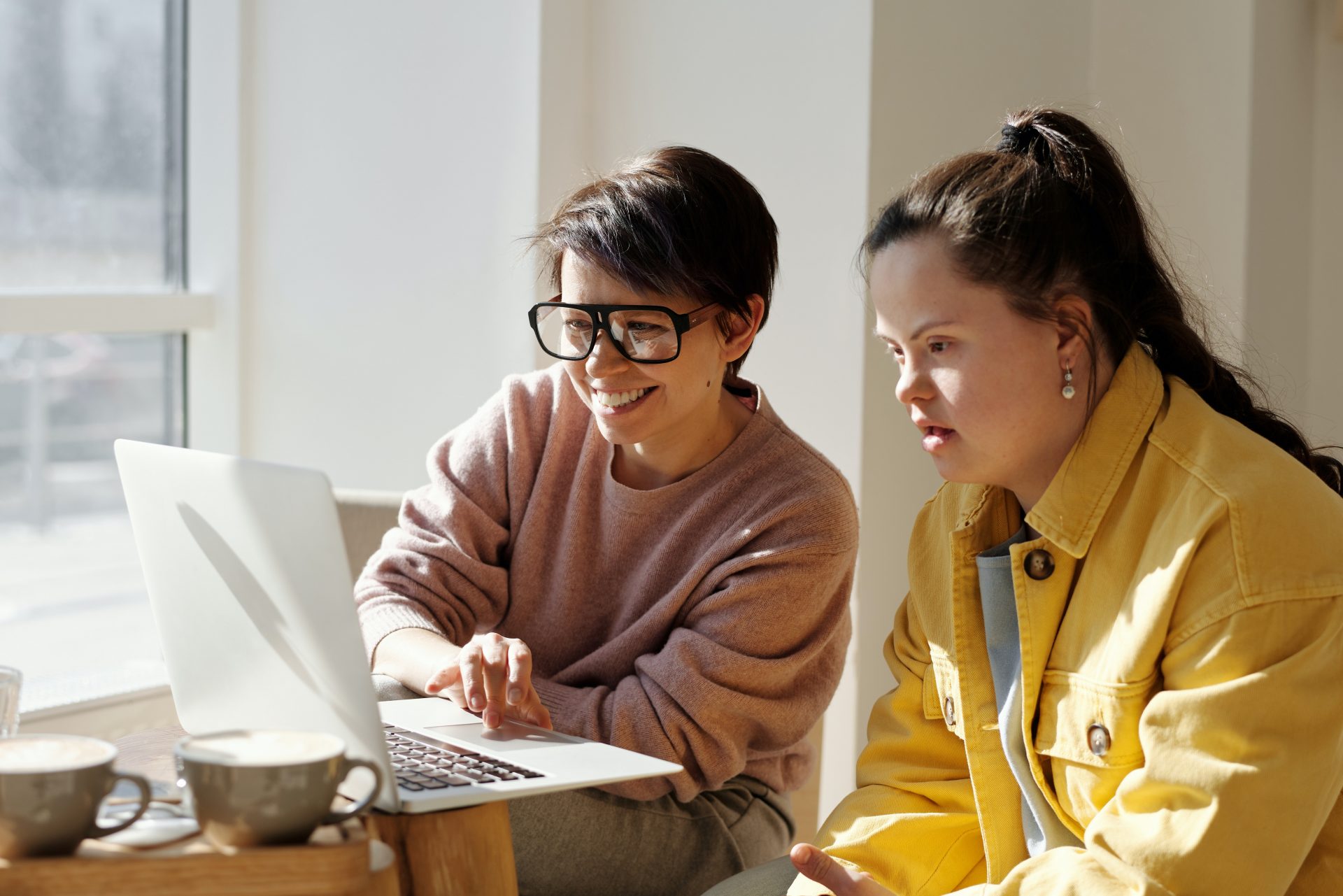 Two women sit at a table looking at the screen of a laptop computer