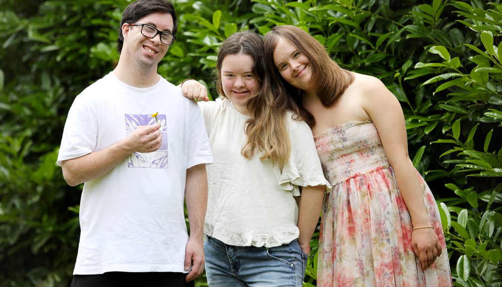 A young man with short dark hair and glasses, and two young women with long dark hair have their arms around each other and are smiling at the camera.