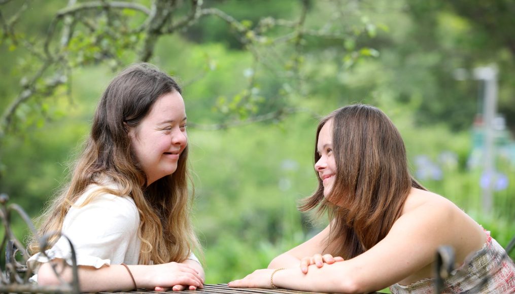 Two young women with long brown hair are sitting at an outdoor table smiling at each other.