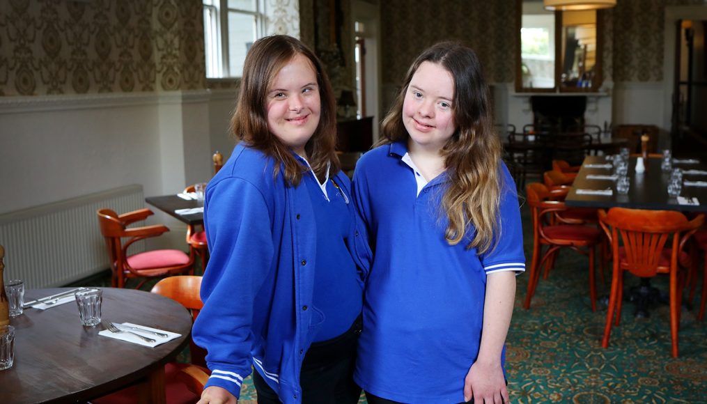 Two young women with long dark hair are wearing blue uniforms and smiling at the camera. They are standing in a dining room.